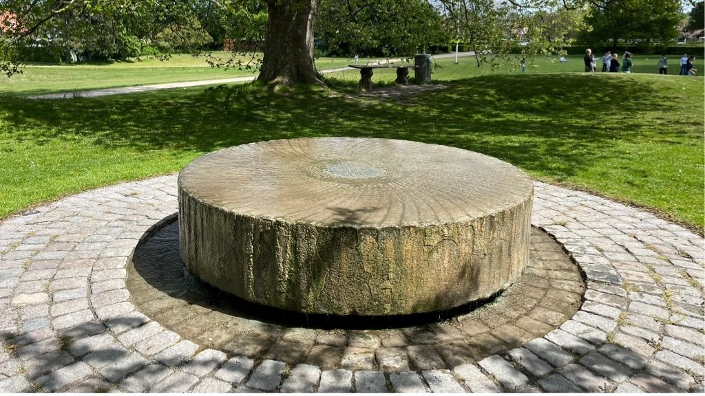 Round stone fountain at the Spring in Roskilde Bypark, surrounded by grass and trees.