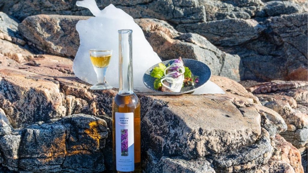 En flaske snaps, et glas og en tallerken mad står på klipper med et stykke is i baggrunden.