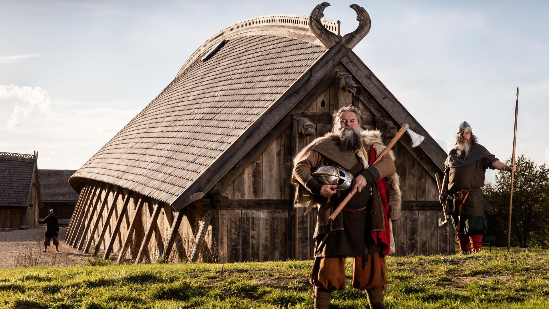 Tre personer i vikingedragter foran Kongehallen i Sagnlandet Lejre – en stor træsalsbygning med udskårne dragehoveder på taget, der gengiver livet i vikingetiden.