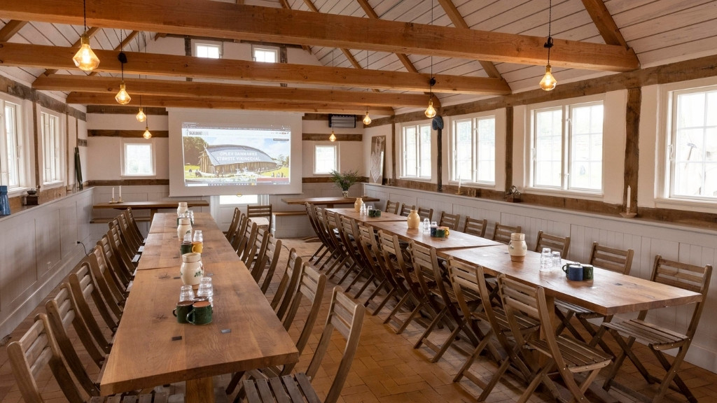 Mødelokale i Sagnlandet Lejre med lange træborde, træstole og projektor.