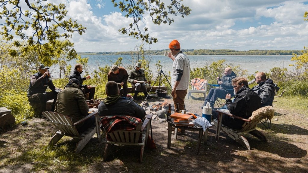 Gruppe nyder fælles måltid ved bål og sø ved Bjælkehytten.