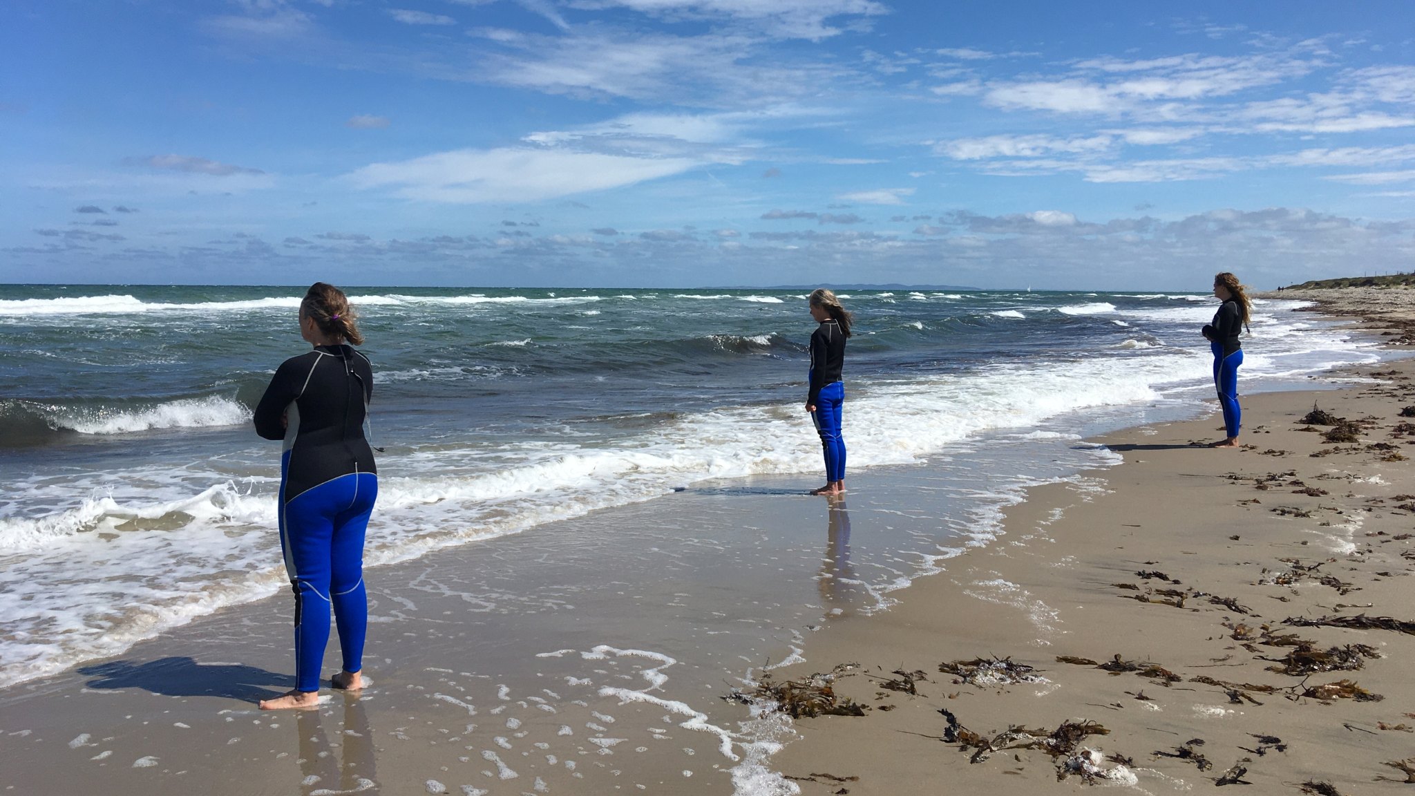 Tre personer i våddragter står ved vandkanten på en sandstrand og kigger ud over havet.