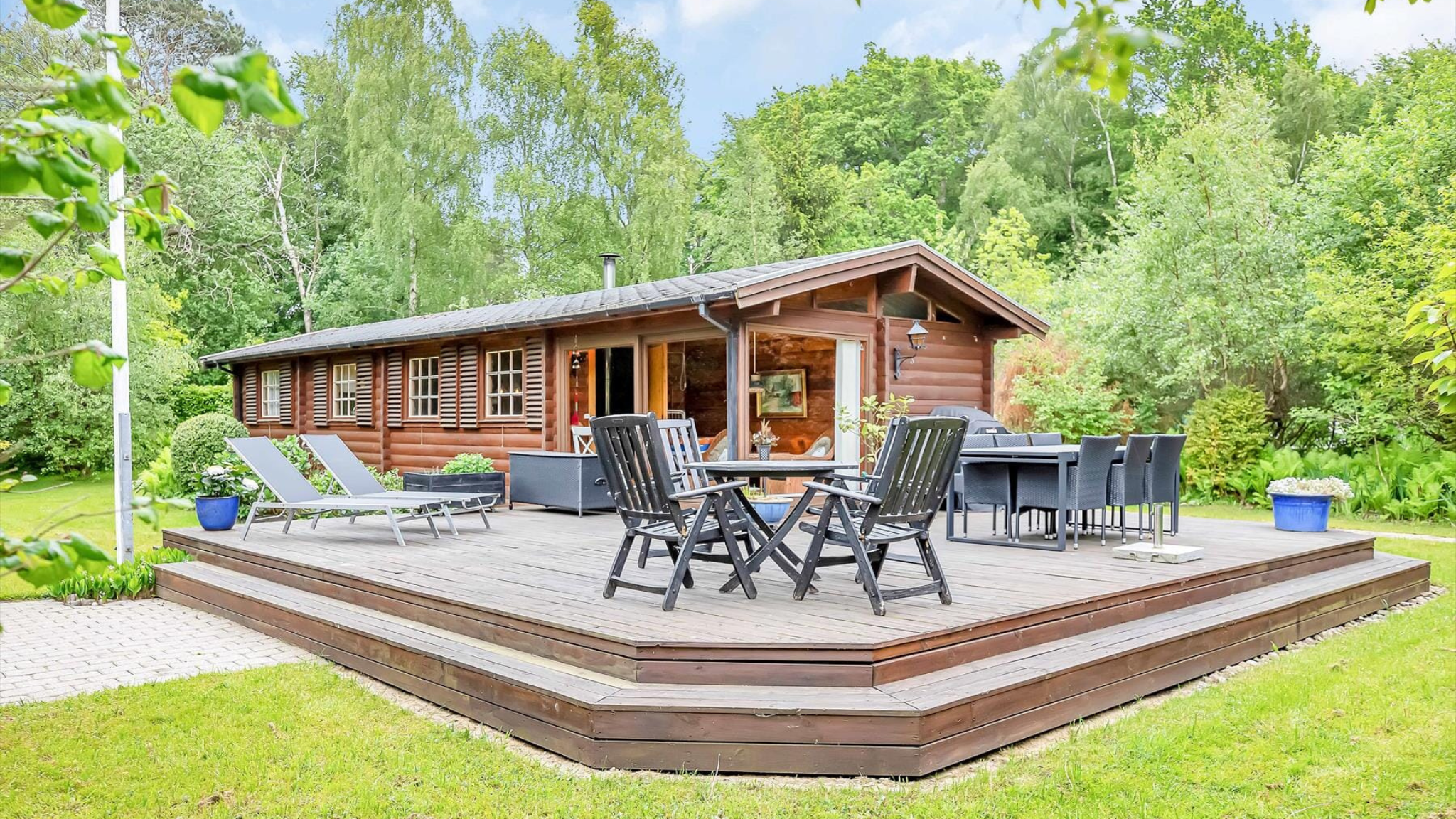 Sommerhus med stor træterrasse, havemøbler og grønne omgivelser i Fjordlandet.