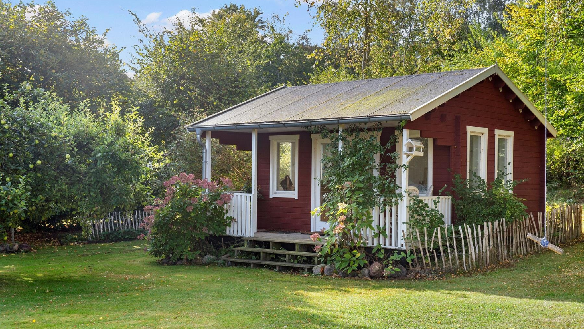 Rødt sommerhus omgivet af grøn have og blomster.