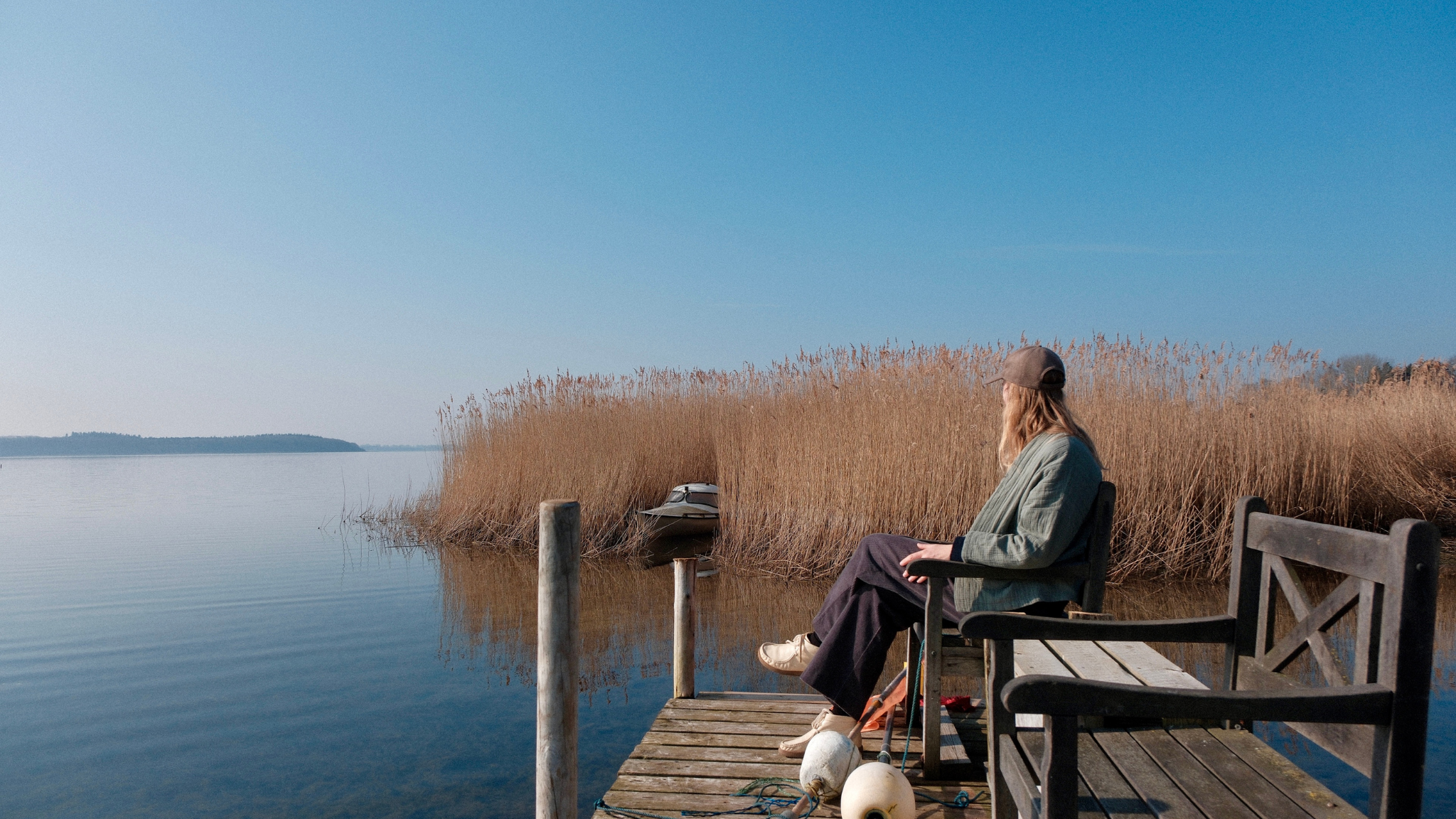 Person sitzt auf einem Holzsteg an einem ruhigen See, umgeben von Schilf unter blauem Himmel.