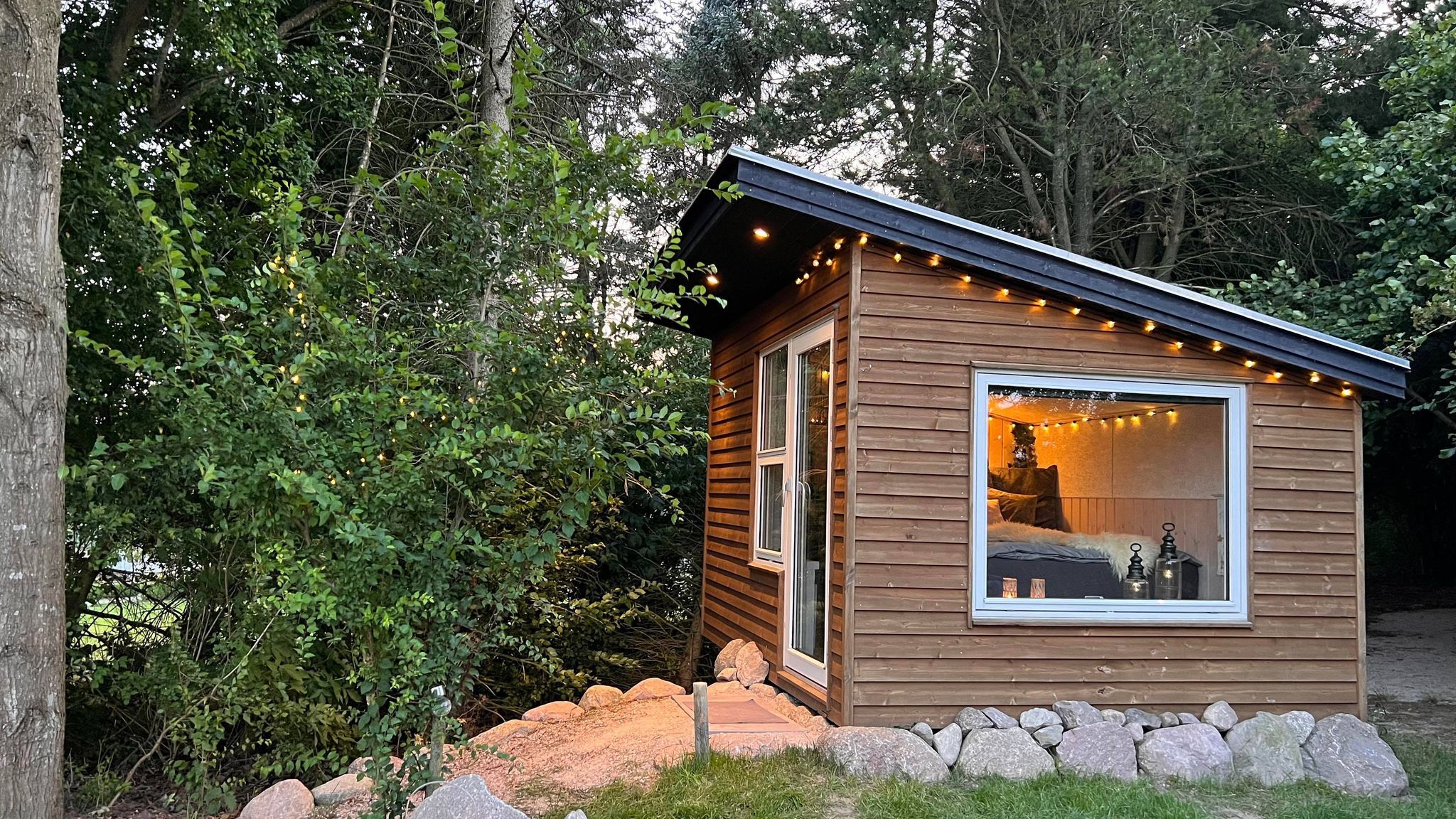 Tiny House von Heart of Nature, umgeben von Bäumen, mit stimmungsvoller Beleuchtung und großen Fenstern.