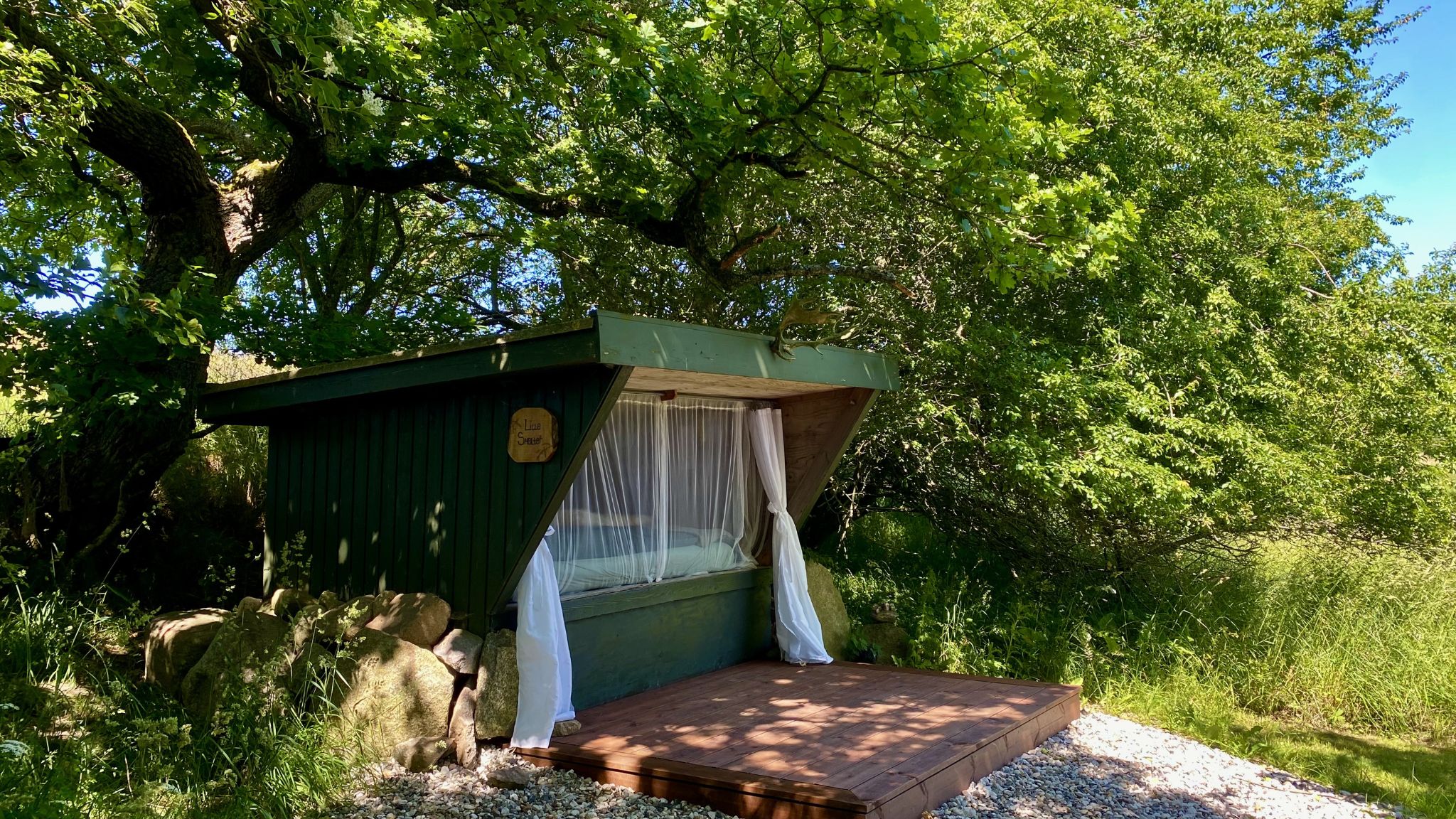 Shelter von Heart of Nature unter großen Bäumen mit offenem Schlafbereich und Blick in die Natur.
