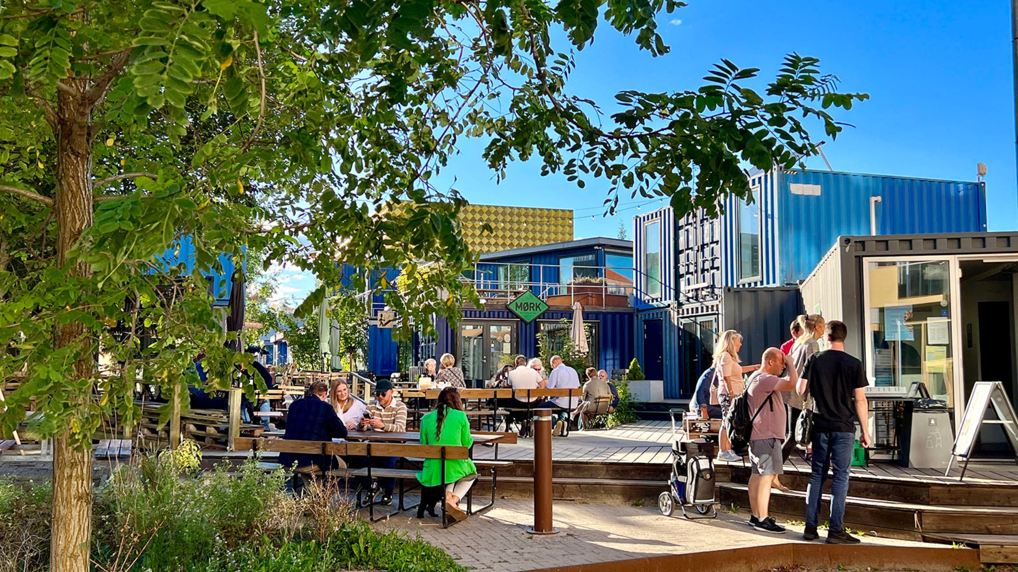People at outdoor tables between containers and trees at Musicon