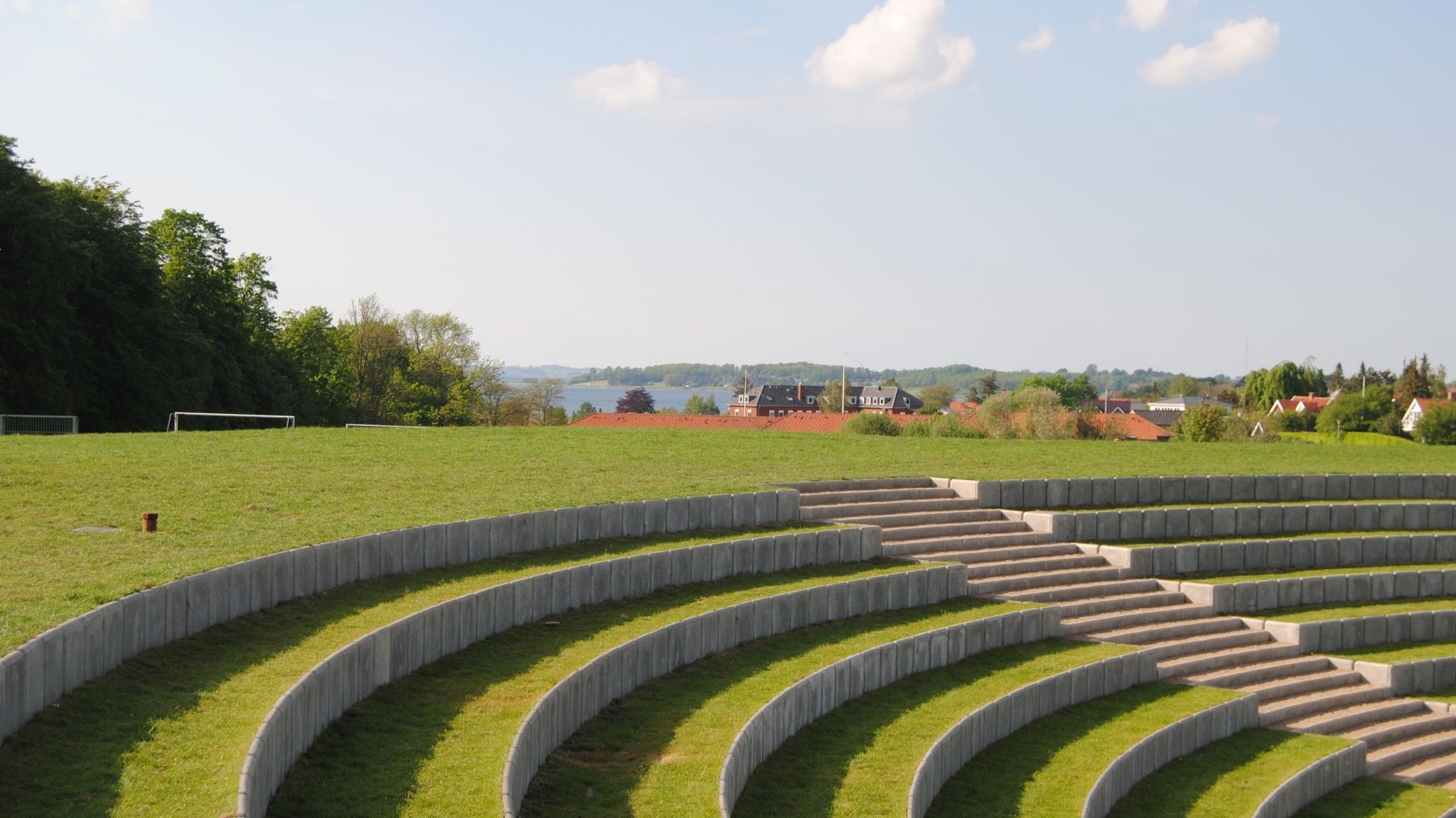 Outdoor amphitheatre with grass-covered seating and views of houses, trees and water.