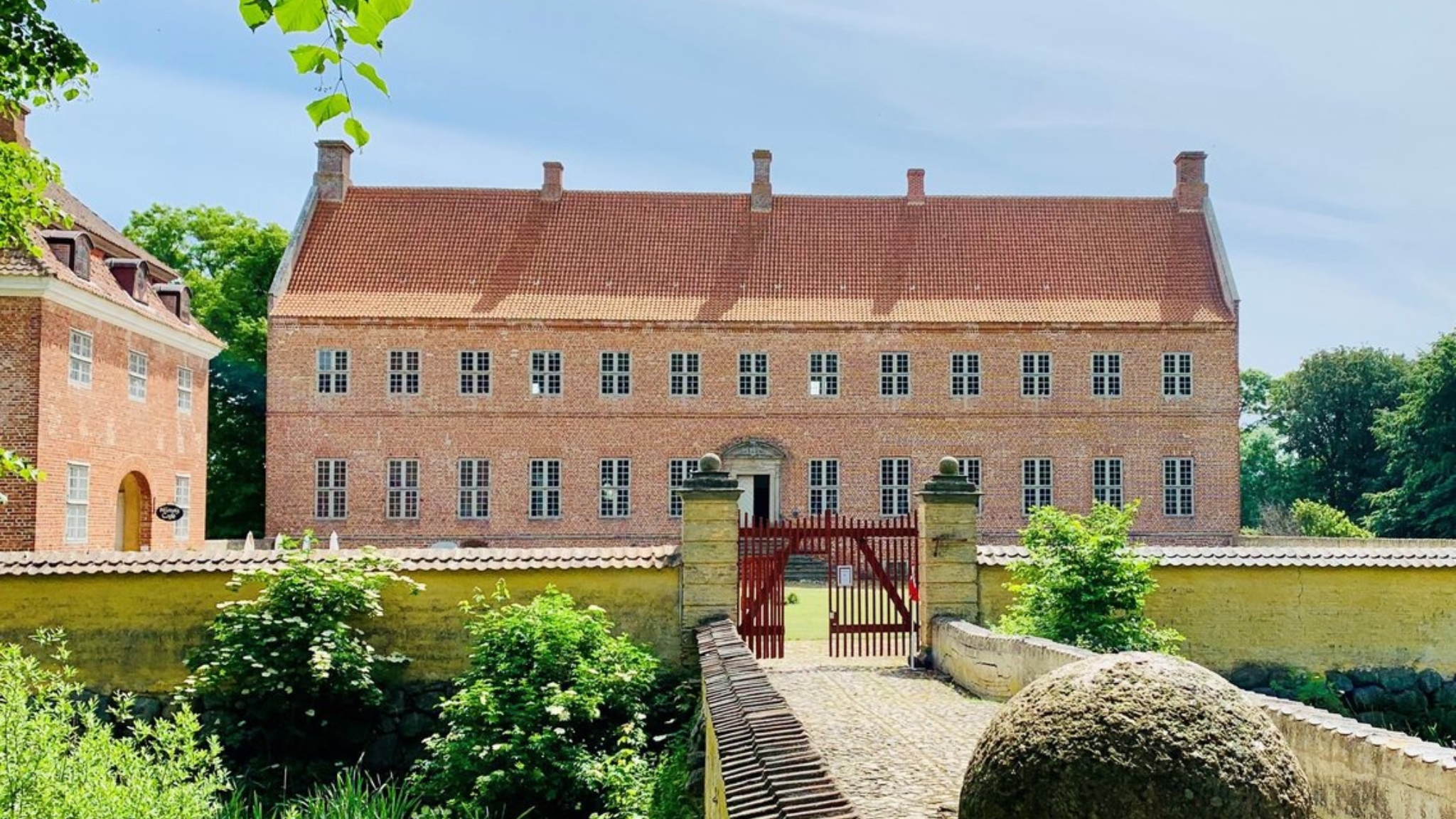 Schloss Selsø mit historischem Hauptgebäude, Tor und Brücke, umgeben von grünen Bäumen.