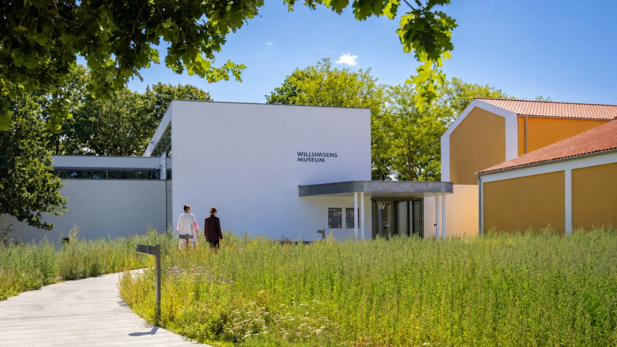 Willumsens Museum with white and yellow buildings surrounded by green vegetation and trees.