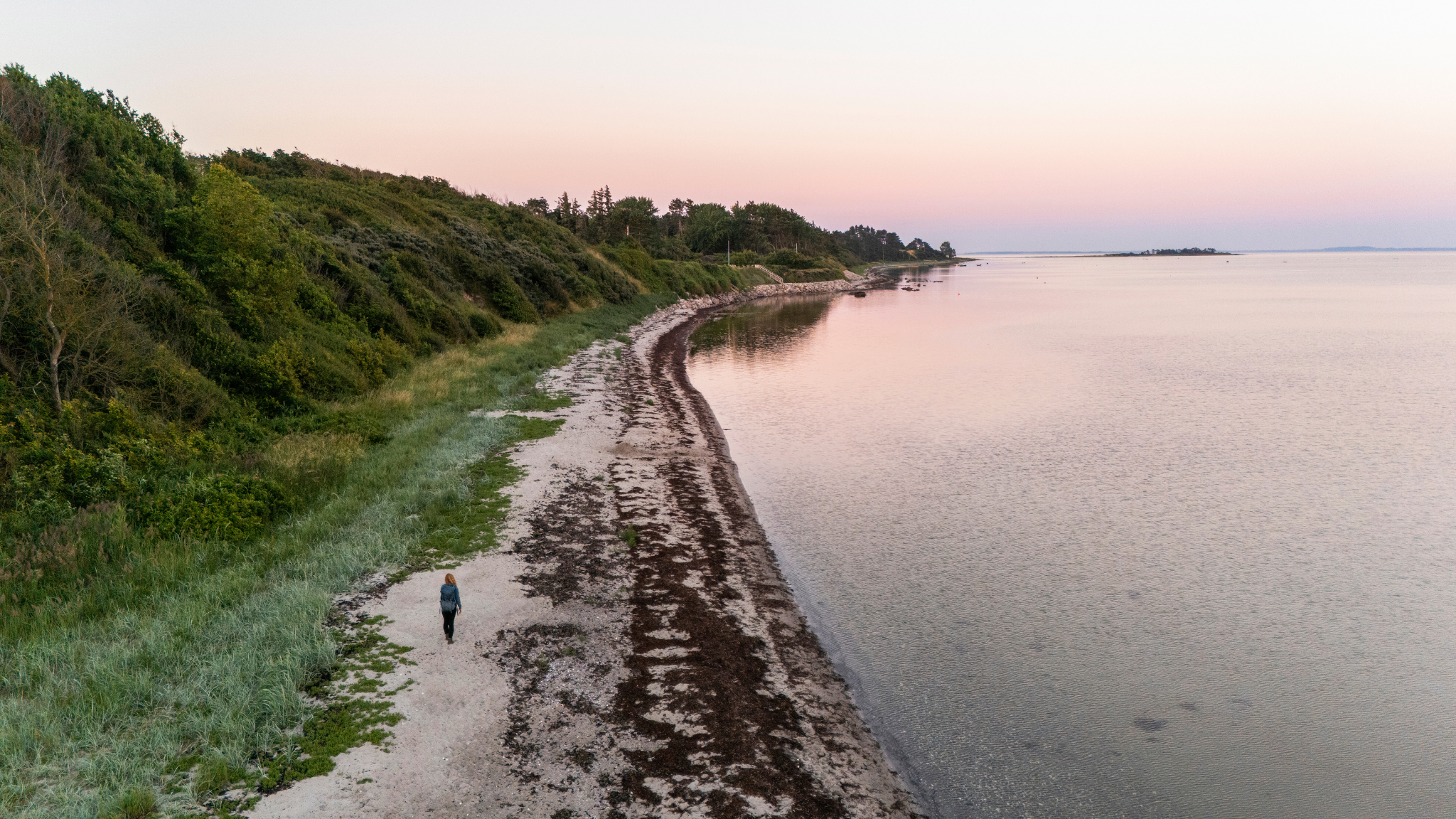 Person walking along Kulhuse Strand with a green coastline and calm water in the evening light.