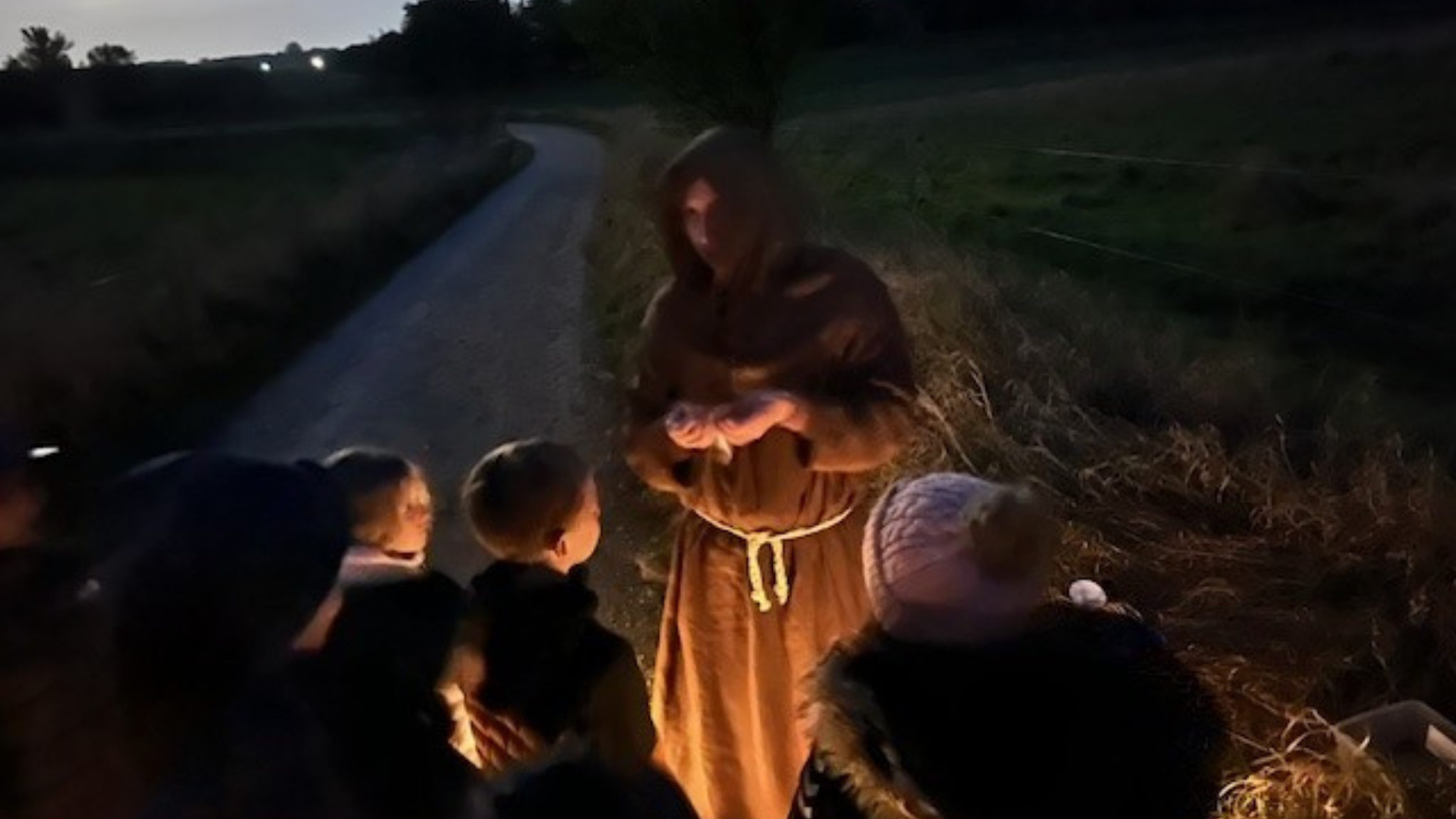 Historically dressed guide telling stories to children by a country road after dark.
