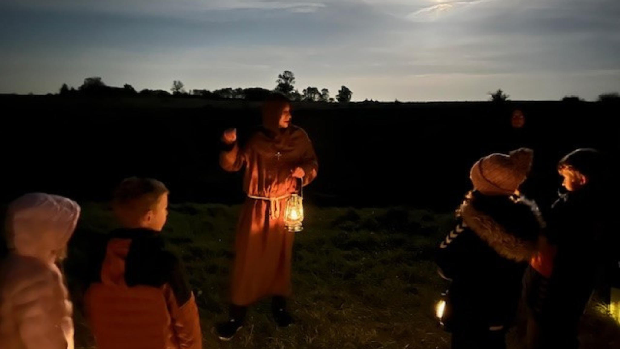Guide in historical costume holding a lantern while telling children stories outdoors in the evening.