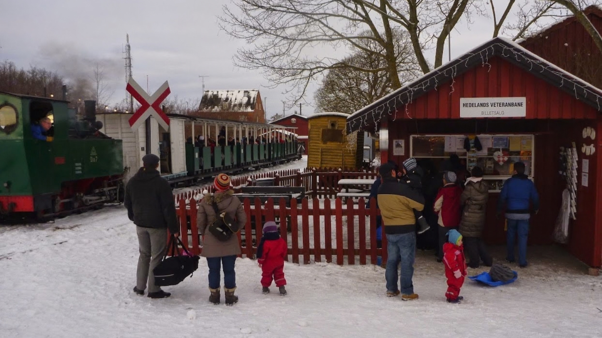 Kinder und Erwachsene am Fahrkartenverkauf von Hedelands Veteranbane mit Dampfzug und Schnee im Hintergrund.