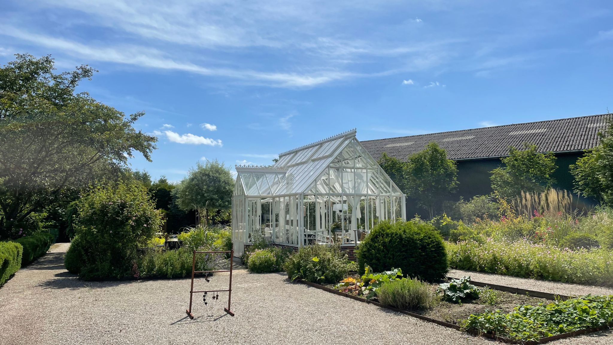 White greenhouse surrounded by flower beds, green shrubs and gravel paths in a lush garden.