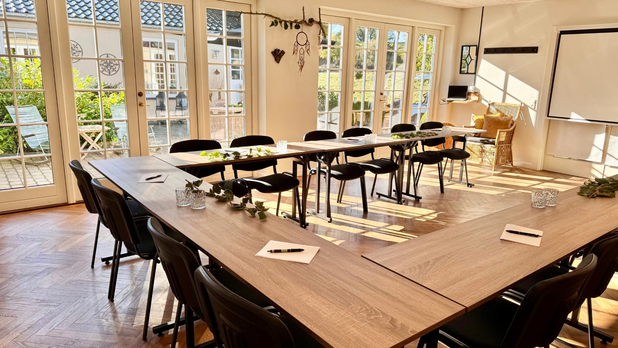 Bright meeting room with U-shaped tables, black chairs, large windows and a presentation screen.
