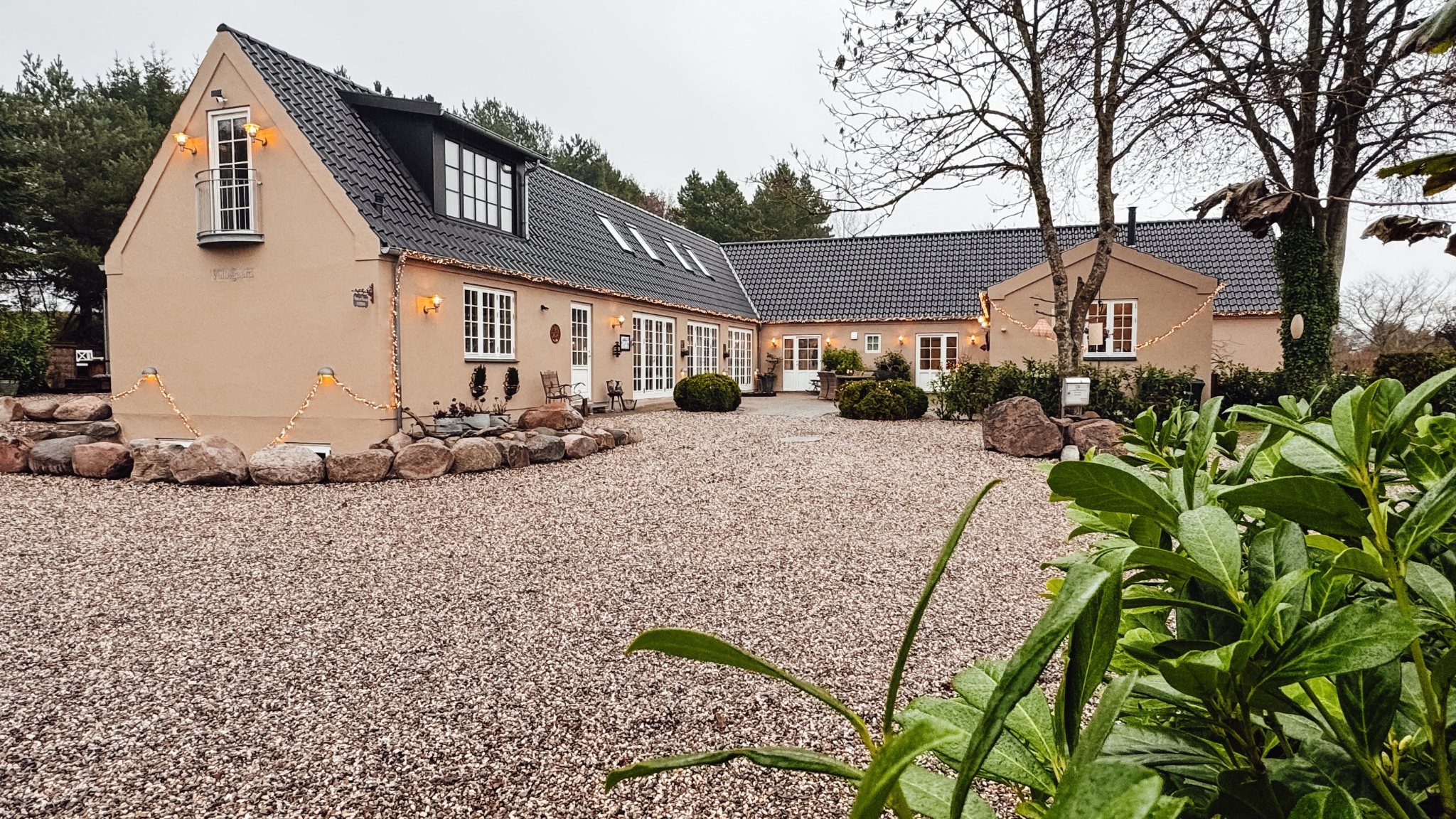 Light-coloured country property with a black tiled roof, gravel courtyard and atmospheric outdoor lighting.