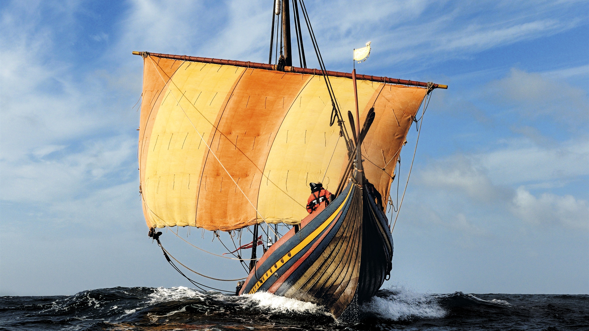 Træskib set forfra med gult råsejl på vandet under en blå himmel.
