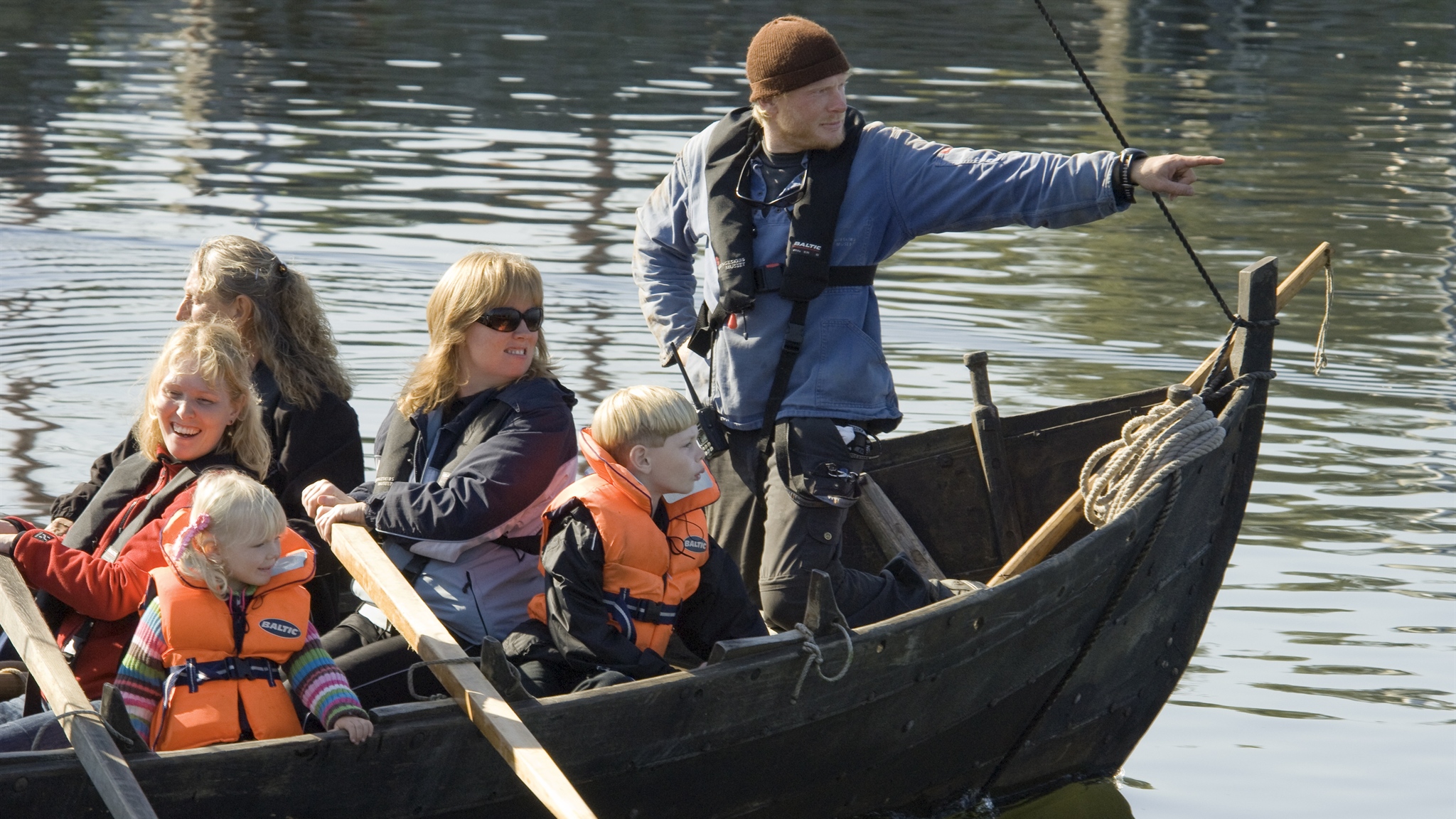 Voksne og børn med redningsveste i en træbåd med en instruktør fra Vikingeskibsmuseet.