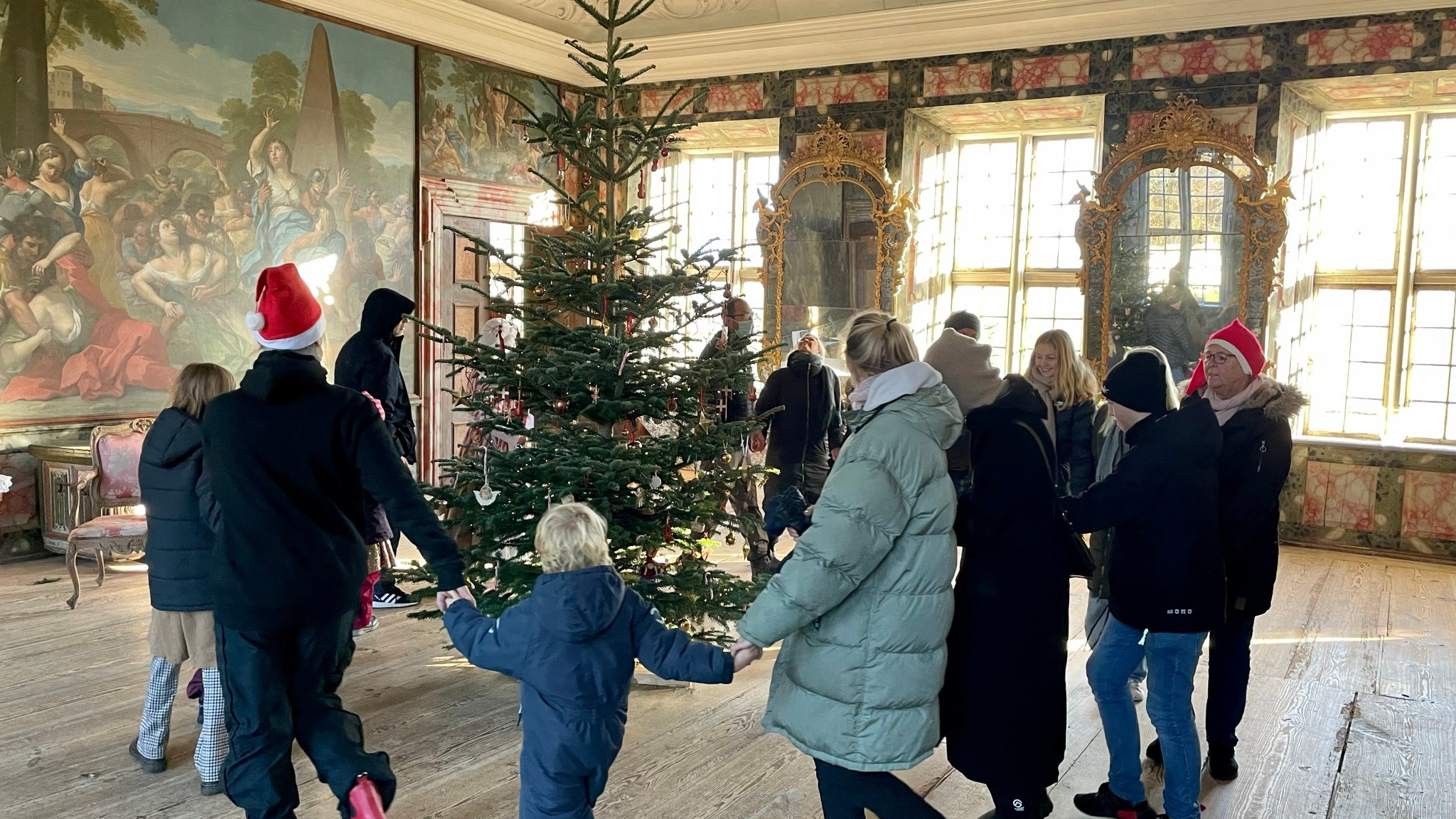 Børn og voksne danser om juletræet i en historisk sal på Selsø Slot under julearrangement.