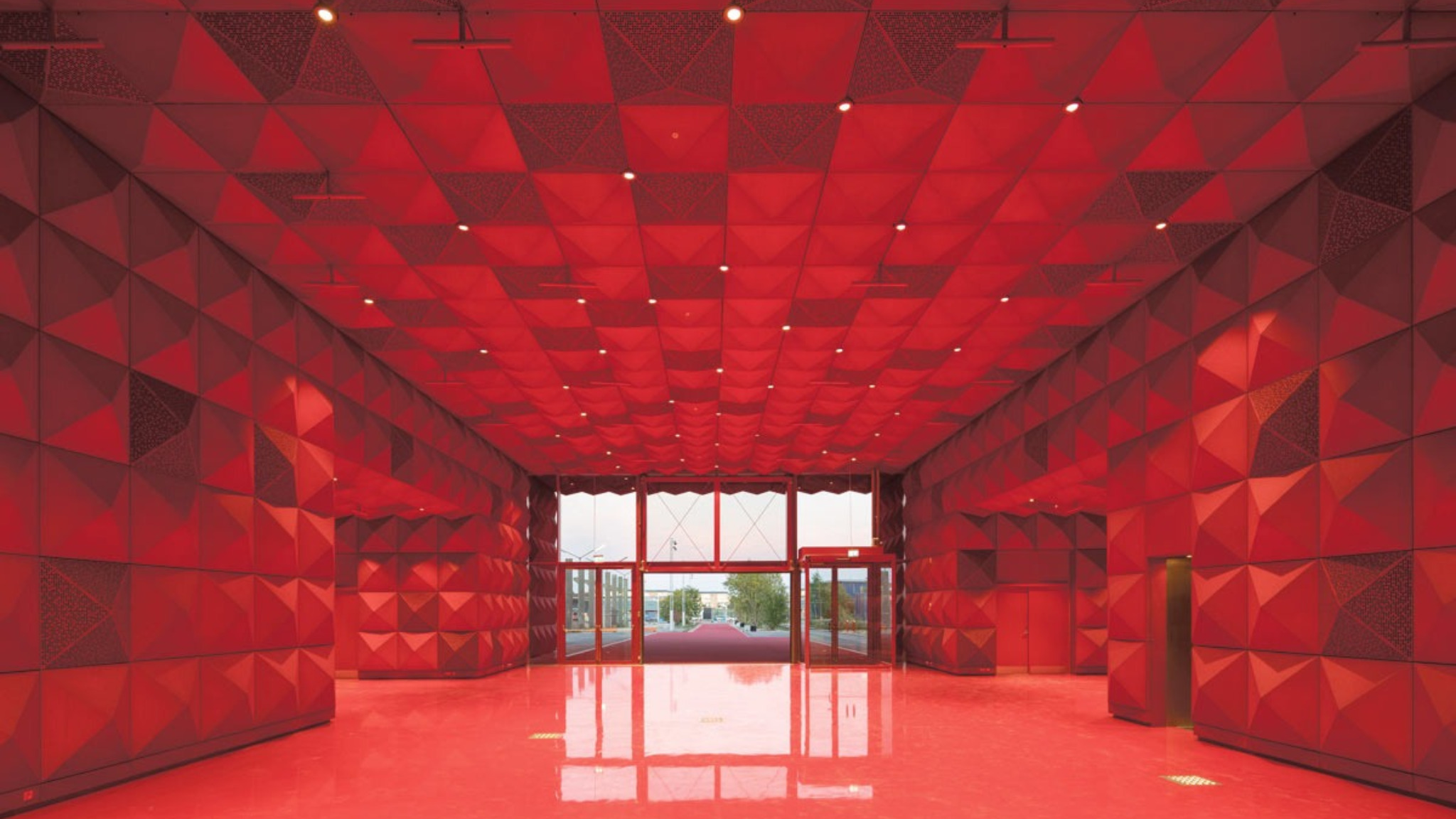 Rotes Foyer im Rockmuseum RAGNAROCK in Roskilde mit geometrischen Wandpaneelen, hoher Decke und Glaseingang.
