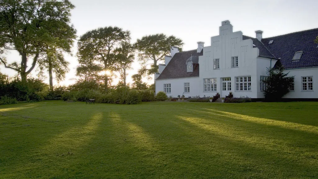 Brandbjerg Manor's facade with light falling through the trees on a beautifully freshly mowed green lawn.