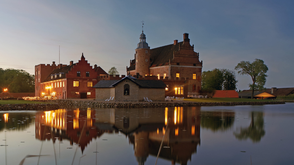 Stort rødt slot ses ved aftentid med tændte lys i vinduerne, der spejler sig flot i vandet.