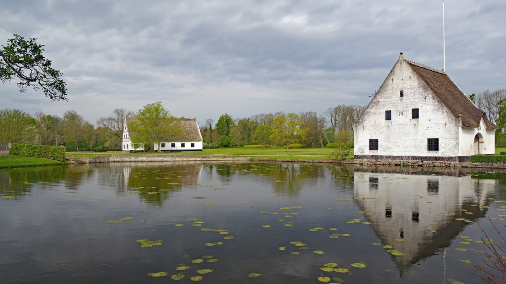 Hvidt gods med mørkt tag ligger og spejler sig i noget vand med grønne omgivelser.