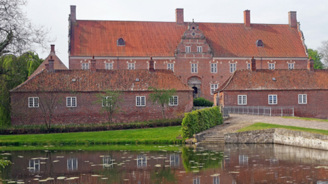 Stort rødt gods med rødt tag ligger på en grøn plæne med noget vand i forgrunden.