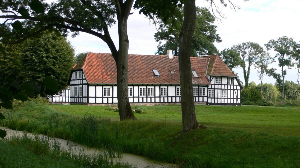 Hvid herregård i bindingsværk med rødt tag set bag nogle træer.