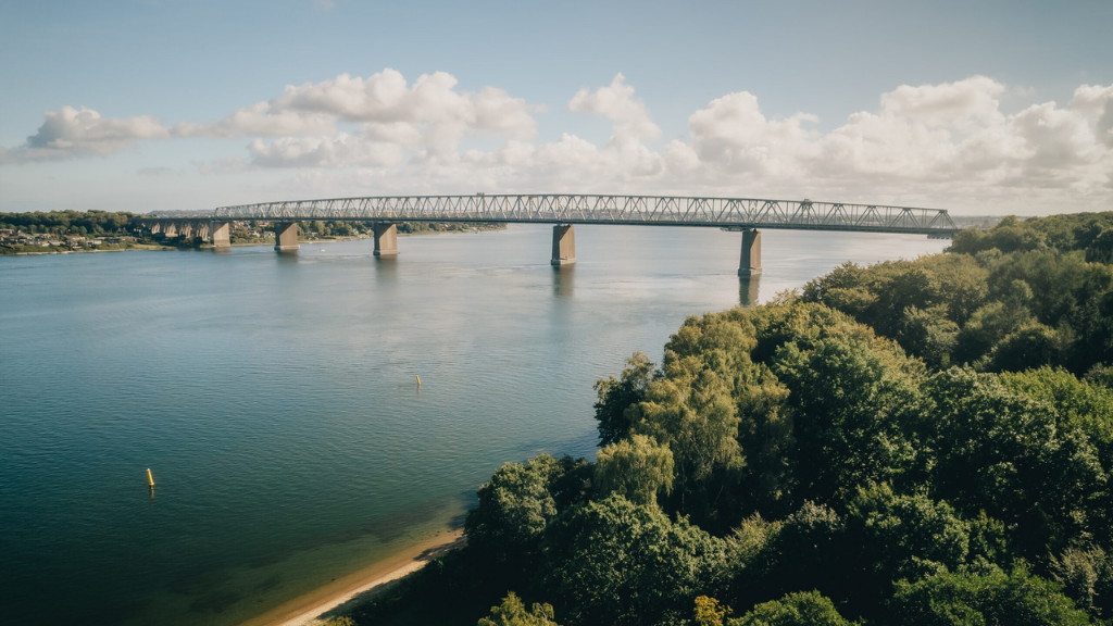 Droneshot af en bro, der fører på tværs af et flot, blåt hav med grøn skov til højre.
