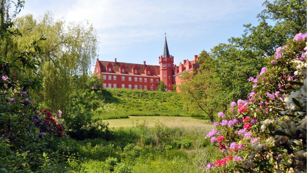 Rotes Herrenhaus mit Turm, aus der Ferne durch einen wunderschönen, farbenfrohen Garten gesehen.